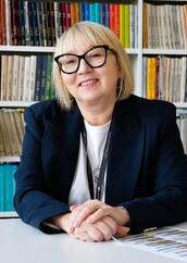 Kobieta w średnim wieku z blond włosami i czarnymi okularami siedzi przy białym stole w bibliotece. Ma na sobie granatową marynarkę i biały t-shirt, a na szyi zawieszony identyfikator. Uśmiecha się serdecznie, spoglądając w stronę obiektywu. W tle znajdują się regały wypełnione książkami w kolorowych okładkach. Na stole leży dokument z tekstem.