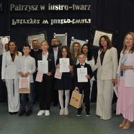 Uczniowie z dyplomami i dorośli stoją razem na scenie z dekoracją lustrzaną i napisem „Patrzysz w lustro – twarz”.