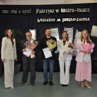 Uhonorowani uczestnicy z kwiatami i dyplomami stoją na scenie przed dekoracją z lustrami.
