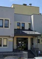 Widok frontowy budynku Filii nr 9 Książnicy Podlaskiej przy ul. Gajowej 73 w Białymstoku, uchwycony od strony ulicy. Dwupiętrowy budynek z jasną elewacją i napisem „BIBLIOTEKA" w prawej części fasady. Widoczne wejście główne z zadaszeniem, schody oraz oznaczenie adresowe „73". Po prawej stronie drzewo liściaste. Na pierwszym planie fragment jezdni z zaparkowanymi samochodami. Zdjęcie wykonane w słoneczny dzień.