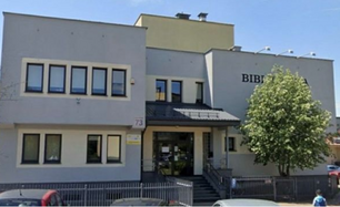 Widok frontowy budynku Filii nr 9 Książnicy Podlaskiej przy ul. Gajowej 73 w Białymstoku, uchwycony od strony ulicy. Dwupiętrowy budynek z jasną elewacją i napisem „BIBLIOTEKA" w prawej części fasady. Widoczne wejście główne z zadaszeniem, schody oraz oznaczenie adresowe „73". Po prawej stronie drzewo liściaste. Na pierwszym planie fragment jezdni z zaparkowanymi samochodami. Zdjęcie wykonane w słoneczny dzień.