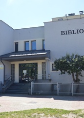 Nowoczesny budynek biblioteki z jasną elewacją, z dużym czarnym napisem "BIBLIOTEKA" na frontowej ścianie. Wejście z przeszklonymi drzwiami i schodami, po bokach metalowe ogrodzenie, przed budynkiem drzewko i kostka brukowa. Numer budynku "73" widoczny na fasadzie.