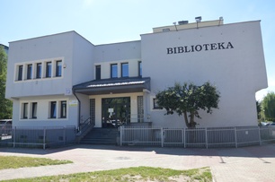 Nowoczesny budynek biblioteki z jasną elewacją, z dużym czarnym napisem "BIBLIOTEKA" na frontowej ścianie. Wejście z przeszklonymi drzwiami i schodami, po bokach metalowe ogrodzenie, przed budynkiem drzewko i kostka brukowa. Numer budynku "73" widoczny na fasadzie.