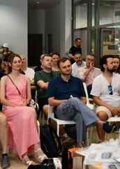 Grupa ludzi w różnym wieku siedzi na krzesłach w nowoczesnym, jasnym pomieszczeniu, uważnie słuchając prezentacji. Niektórzy mają na sobie letnie ubrania i okulary przeciwsłoneczne, a na stoliku z przodu widać otwartą książkę oraz roślinę w doniczce.