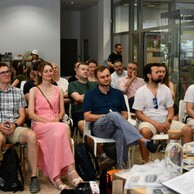 Grupa ludzi w różnym wieku siedzi na krzesłach w nowoczesnym, jasnym pomieszczeniu, uważnie słuchając prezentacji. Niektórzy mają na sobie letnie ubrania i okulary przeciwsłoneczne, a na stoliku z przodu widać otwartą książkę oraz roślinę w doniczce.