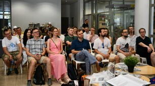 Grupa ludzi w różnym wieku siedzi na krzesłach w nowoczesnym, jasnym pomieszczeniu, uważnie słuchając prezentacji. Niektórzy mają na sobie letnie ubrania i okulary przeciwsłoneczne, a na stoliku z przodu widać otwartą książkę oraz roślinę w doniczce.