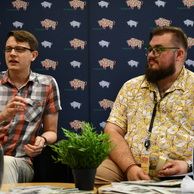 Dwóch mężczyzn siedzi obok siebie podczas panelu dyskusyjnego na tle ciemnoniebieskiej ścianki z kolorowym logo przedstawiającym żubra. Mężczyzna po lewej w koszuli w kratę mówi i gestykuluje, podczas gdy mężczyzna po prawej w żółtej koszuli w kwiaty słucha z uśmiechem.