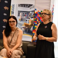 Dwie kobiety podczas oficjalnego wydarzenia. Jedna siedzi na ciemnej kanapie w beżowej sukience, a druga stoi obok niej w czarnej sukience i okularach z grubymi oprawkami, przemawiając do zgromadzonych. W tle widać baner ze zdjęciem nowoczesnej biblioteki.