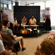 Widok z perspektywy publiczności na salę spotkań, gdzie na niskim podwyższeniu trwa panel dyskusyjny. Trzech mężczyzn siedzi na czarnej kanapie na tle ścianki z napisem „Województwo Podlaskie”, obok stoi kobieta w czarnej sukience, a jedna z osób na widowni podnosi rękę.