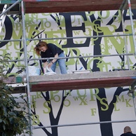 Artystka stoi na rusztowaniach i maluje mural.
