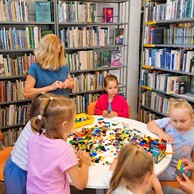 Zdjęcie nr 5 przedstawia bibliotekarkę w niebieskiej bluzce, która pomaga dzieciom w budowaniu sortowni z klocków LEFO. Dzieci siedzą przy okrągłym, białym stole, w tle są regały z książkami.