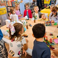 Zdjęcie nr 4 przedstawia dzieci stojące wokół makiety miasta i opowiadające o zbudowanych przez siebie elementach z klocków LEGO. W tle znajdują się regały z książkami.