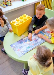 Zdjęcie nr 3 przedstawia dzieci siedzące przy okrągłym zielonym stoliku i oglądające książkę o recyklingu. Na stoliku jest też żółte pudełko w kształcie klocka LEGO.