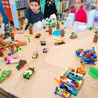 Zdjęcie nr 4  i 8 przedstawia dzieci stojące wokół makiety miasta i opowiadające o zbudowanych przez siebie elementach z klocków LEGO. W tle znajdują się regały z książkami.