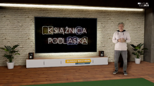 Mężczyzna stoi w wirtualnym studiu telewizyjnym przed dużym ekranem. Ekran wyświetla napis neonowy KSIĄŻNICA PODLASKA. Mężczyzna ma jasny sweter z kapturem (bluza) i szare spodnie dżinsowe. Stoi po prawej stronie ekranu, z rękami złożonymi na wysokości pasa. Pod ekranem znajduje się szafka RTV z głośnikami po bokach, a na niej widoczna jest plakietka z napisem ELIGIUSZ BUCZYŃSKI i pod spodem mniejszymi literami CZYTELNIA OGÓLNA. Ściana za ekranem wygląda jak ściana z czerwonej cegły. Po obu stronach mebla RTV i ekranu znajdują się doniczkowe rośliny. U góry po prawej stronie widać logo z napisem KSIĄŻNICA PODLASKA. Podłoga jest drewniana.