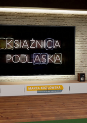 Kobieta stoi w wirtualnym studiu telewizyjnym przed dużym ekranem. Ekran wyświetla podświetlony neonowy napis KSIĄŻNICA PODLASKA. Kobieta ma na sobie czarną bluzkę w różowo-fioletowe kwiaty i niebieskie spodnie dżinsowe. Stoi po prawej stronie ekranu, trzymając ręce swobodnie przed sobą. Pod ekranem znajduje się szafka RTV z głośnikami po bokach, a na niej widoczna jest plakietka z napisem MARTA KOZŁOWSKA i pod spodem mniejszymi literami WYPOŻYCZALNIA GŁÓWNA I MIĘDZYBIBLIOTECZNA. Ściana za ekranem wygląda jak ściana z czerwonej cegły. Po obu stronach mebla RTV i ekranu znajdują się doniczkowe rośliny. U góry po prawej stronie widać logo z napisem KSIĄŻNICA PODLASKA. Podłoga jest drewniana.